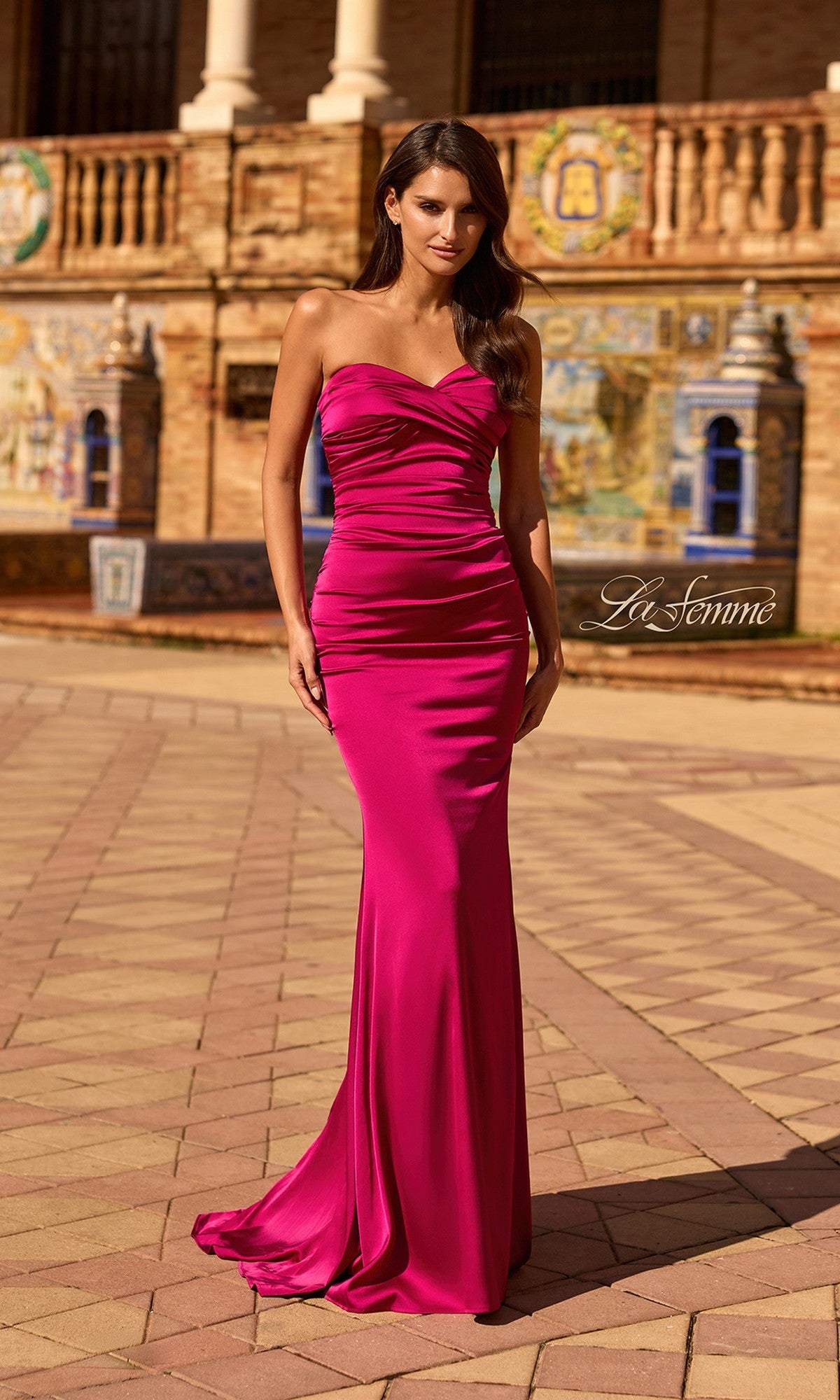 A woman stands outdoors in front of a decorative building wearing the Classic Strapless Long Prom Dress: La Femme 33531, a fitted, floor-length raspberry gown with ruched detailing and a sweetheart neckline.